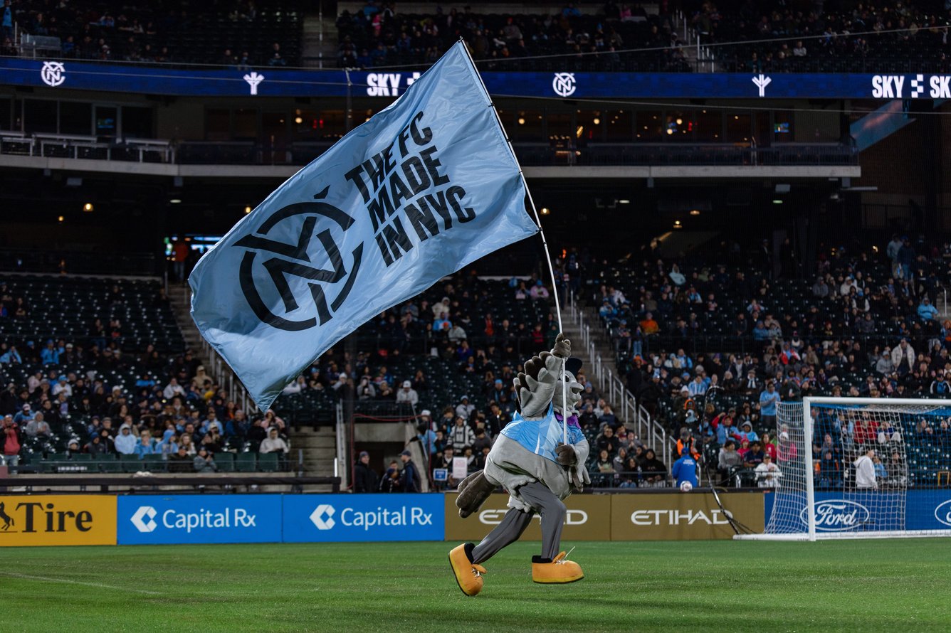 New York City FC mascot Sky Scraper, a pigeon in work books, running on a field carrying a flag.